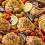Greek sheet pan chicken with roasted zucchini, red onions, cherry tomatoes, and olives, drizzled with olive oil and fresh lemon, served on a baking tray.